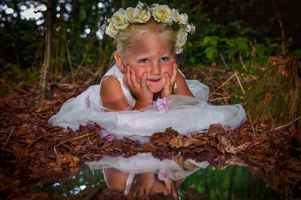 A young girl with a floral wreath sits in a white dress among fallen leaves, resting her chin on her hands, and smiling at the camera with a reflective water surface in front of her.