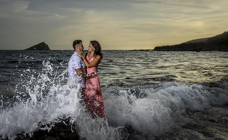 A couple embraces on a rocky beach as waves crash around them, with a calm sea and a distant island in the background under a twilight sky.
