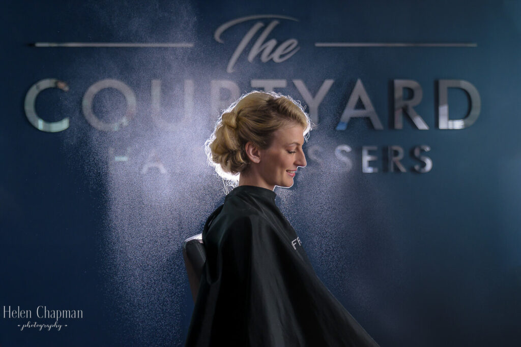A woman in a salon cape sits in front of a sign reading "the courtyard hair raisers," with her blonde hair styled up and a misty ambiance around her.