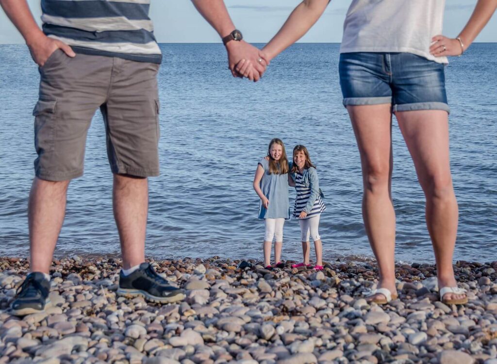 Two children stand holding hands on a pebbled beach, framed by the legs of two adults who are also holding hands, under a clear blue sky.