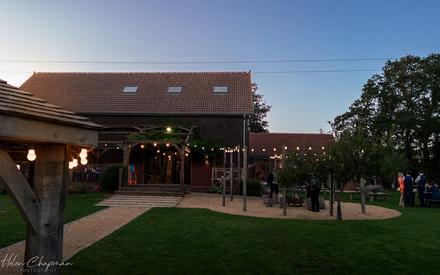 An evening scene at a barn-style venue with warm lighting and guests mingling outside. the barn has a gently lit interior and exterior decorations include string lights and greenery.