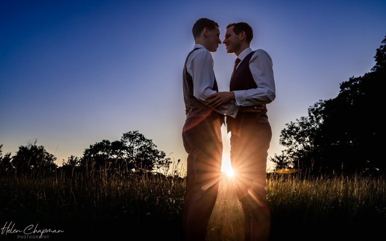 Two men in suits hold hands and gaze at each other in a field at sunset, with the sun casting a dramatic silhouette.