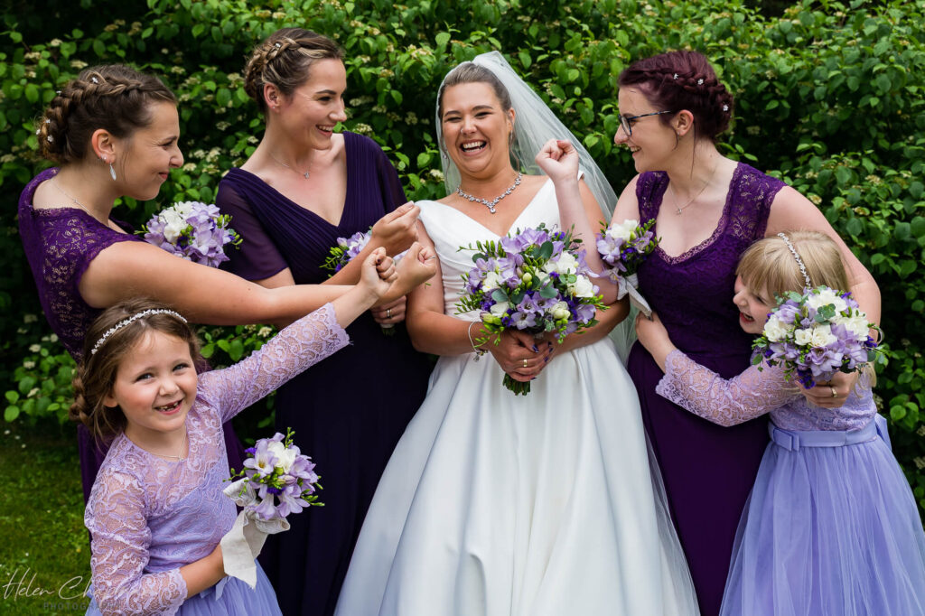 A joyful bride and her bridesmaids, including young girls, laughing and holding hands outdoors. they are dressed in purple and white, holding matching bouquets.
