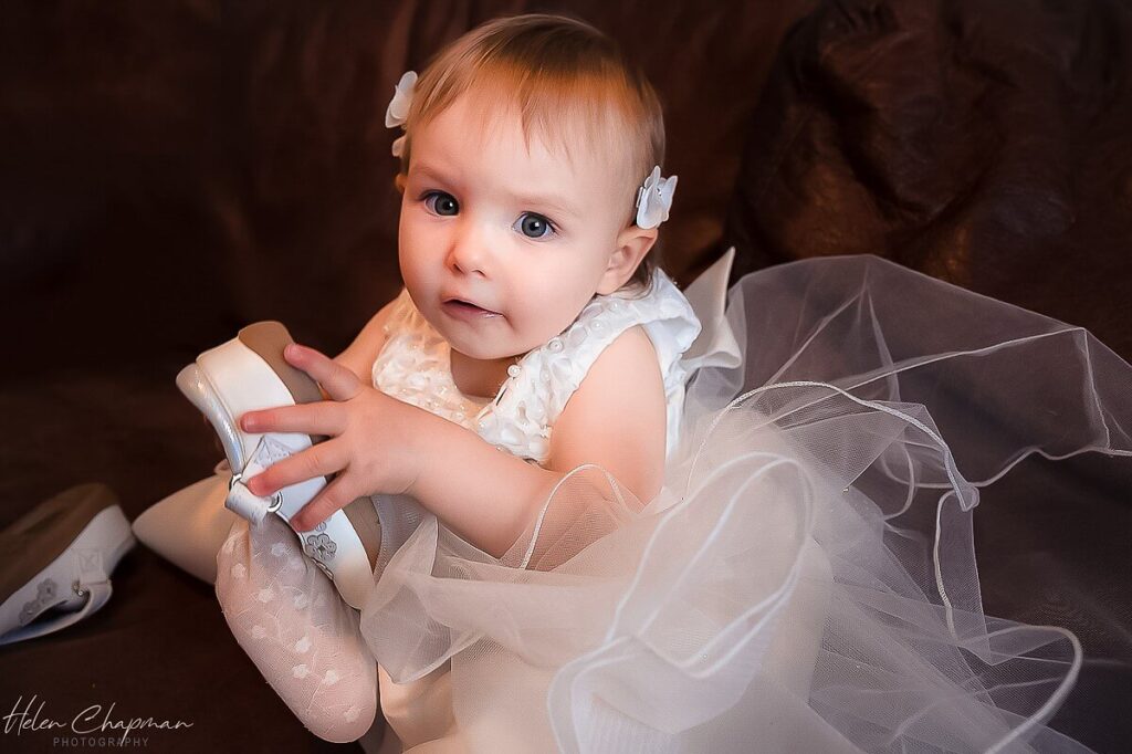 A baby in a white dress with tiny flowers in her hair holds onto a ballet shoe, surrounded by soft tulle fabric, with a curious expression on her face.