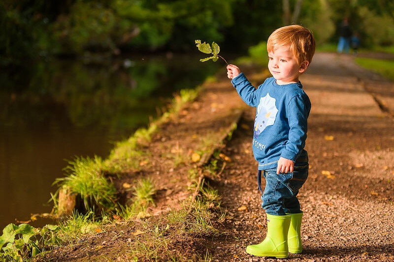 A young child in a blue shirt and green boots holds a leaf, standing by a serene lakeside path, looking thoughtfully in the distance.