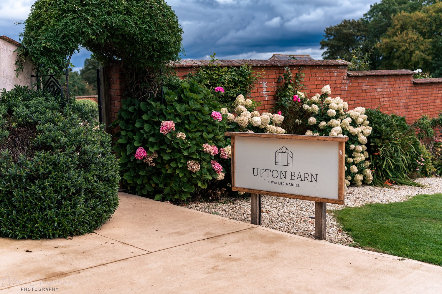 A sign reading "upton barn" in front of a garden with colorful hydrangeas and well-manicured shrubs, set against a rustic brick wall under a cloudy sky.