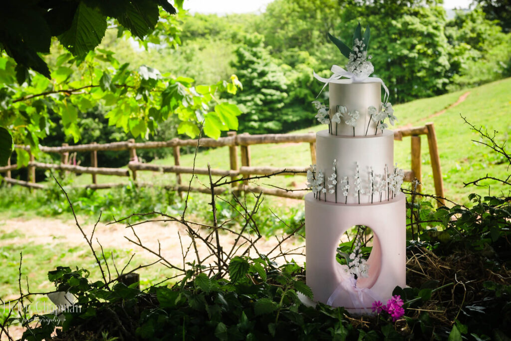 A three-tiered wedding cake decorated with silver and white ornaments, displayed on a table in a lush green garden with a wooden fence in the background.