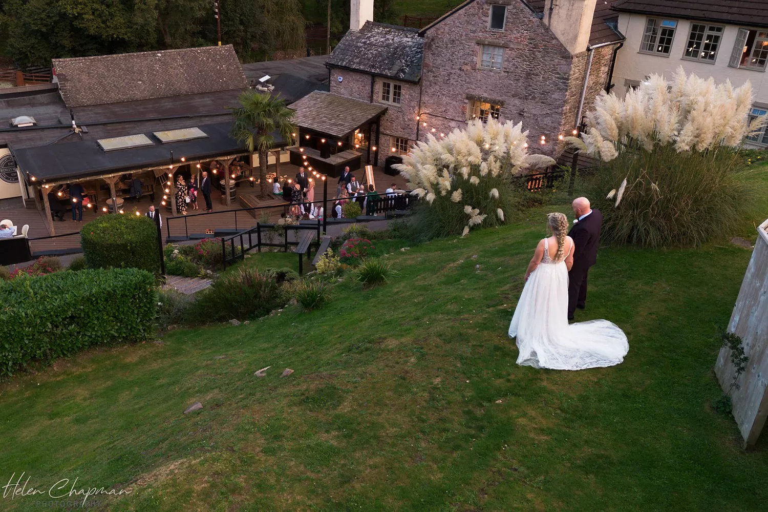 A bride and groom hold hands on a grassy hill, looking down at a warmly lit venue with guests enjoying the evening. a gentle slope leads to the festivities surrounded by stone buildings.