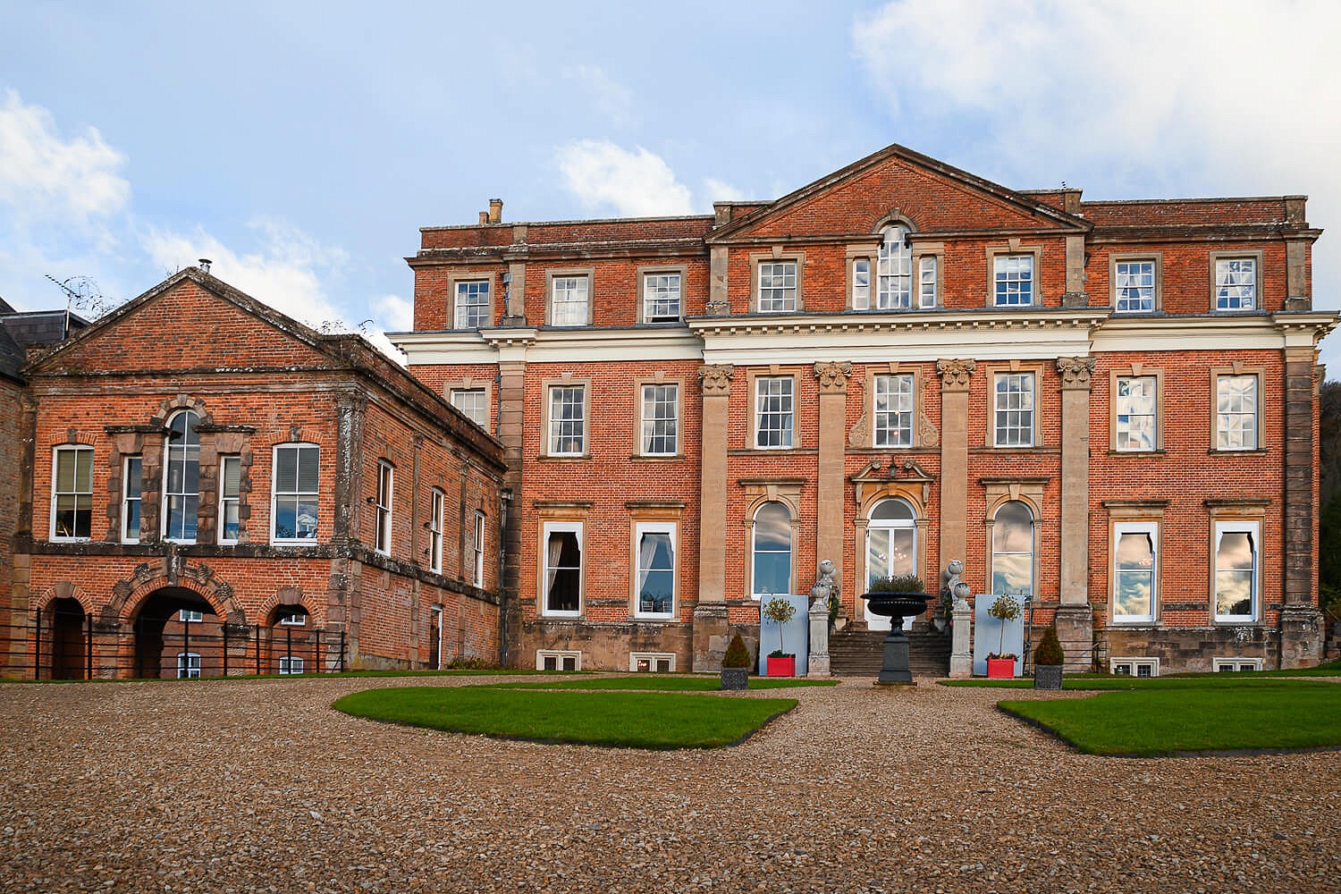A grand historic red brick mansion with evenly spaced white windows, two main doors, and an ornate stone urn in the center, set against a blue sky with clouds, and surrounded by a gravel courtyard.