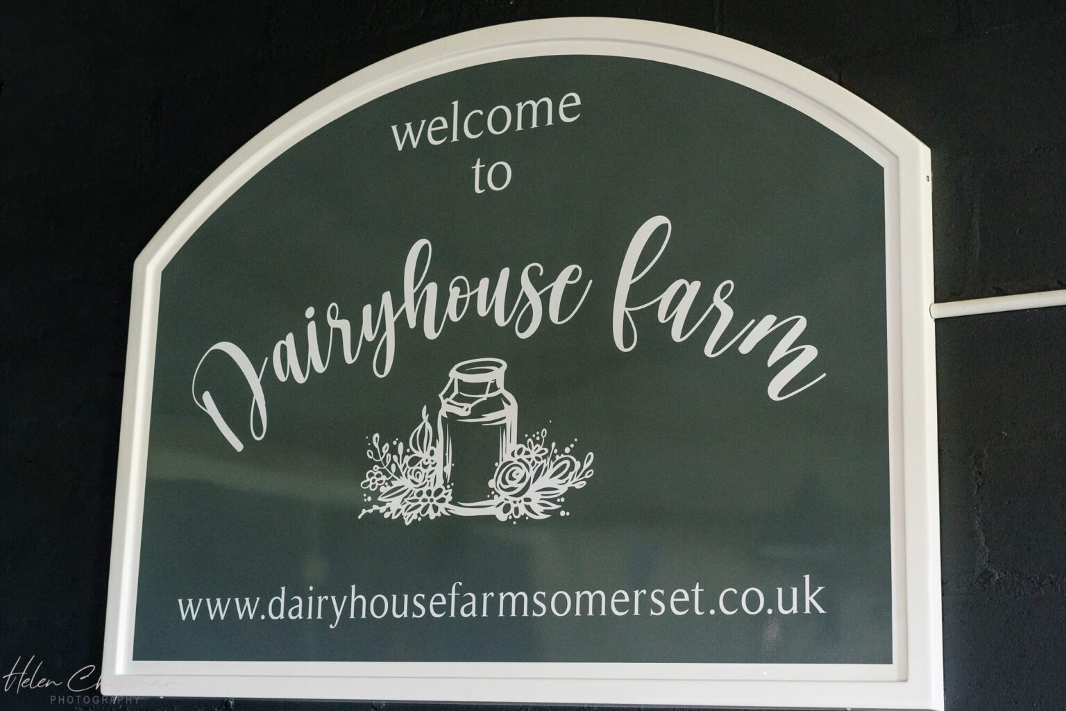 A sign reading "welcome to dairyhouse farm" above the website address "www.dairyhousefarmsomerset.co.uk," elegantly scripted in white on a dark background, with a decorative jar and flowers.