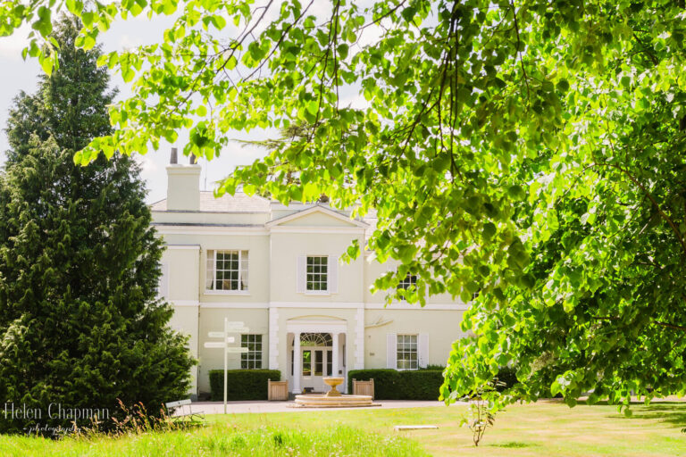 A bright image of a white two-story house with large windows and a central door, set behind a sunlit lawn, framed by overhanging green tree branches.