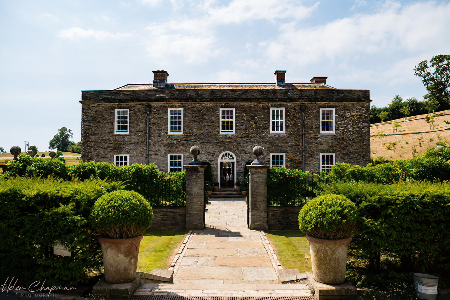 Elegant two-story stone house with a central entrance flanked by round topiary bushes, set against a clear sky and surrounded by well-manicured lawns.