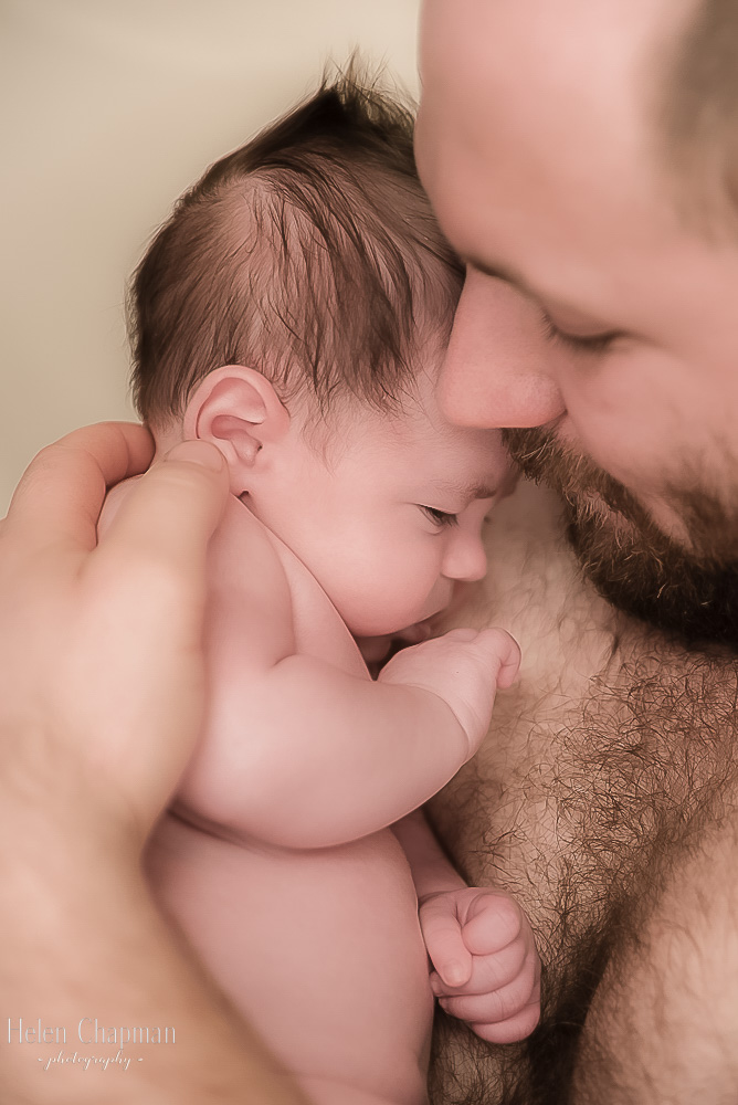 A tender moment between a newborn and a bearded man, where the baby, resting comfortably, touches the man's chest with a tiny hand.